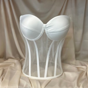 Puede incluir: Un corsé blanco sin tirantes con un corpiño de malla y ballenas. El corsé está sobre un fondo de tela marrón claro.