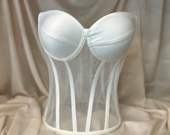 May include: A white strapless corset with a mesh bodice and boning. The corset is on a light brown fabric background.
