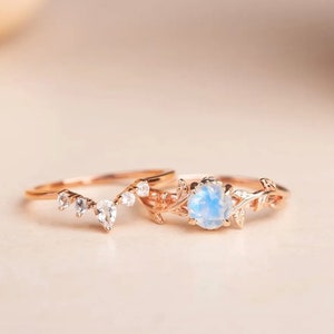 Puede incluir: Dos anillos de oro con delicados diseños florales. El anillo más grande presenta una piedra preciosa redonda de color azul claro. El anillo más pequeño tiene una forma curva con cinco pequeñas piedras preciosas transparentes.