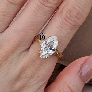 Op de afbeelding: Een gouden ring met een grote, ovaal geslepen, heldere edelsteen die door meerdere pootjes wordt vastgehouden. De ringband bestaat uit verschillende dunne, parallelle gouden draden die samenkomen bij de zetting. Een zwart logo met de letters TD is zichtbaar.