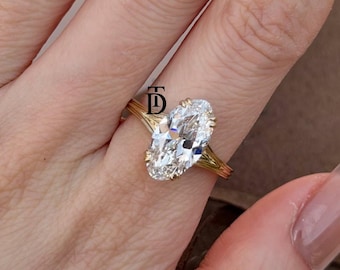 May include: A gold ring featuring a large, oval-cut, clear gemstone held by multiple prongs. The band consists of several thin, parallel gold wires that meet at the setting. A small black logo with the letters TD is visible above the gemstone.