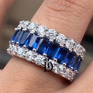 May include: A silver ring featuring a row of rectangular, deep blue gemstones, flanked by rows of smaller, round, clear gemstones. The ring is set on a finger, showcasing its design and sparkle. The ring's design is elegant and eye-catching.