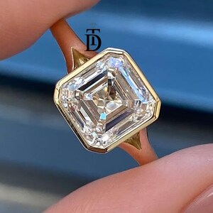 May include: A gold ring featuring a square-cut, bezel-set clear gemstone with a prominent step-cut facet pattern. The band is thin and polished, and a small logo with the letter D is visible on the metal setting near the stone's edge.