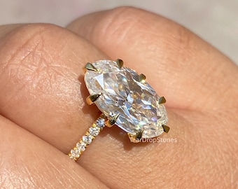 May include: A gold ring featuring a large oval-cut clear gemstone held by eight prongs. The band is thin and set with small round clear stones along the side. The text TearDropStones is visible near the base of the setting.