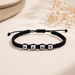 Könnte beinhalten: Ein schwarzes Armband aus geflochtener Kordel mit vier weißen quadratischen Perlen, die mit schwarzen Buchstaben das Wort NAME bilden. Das Armband verfügt über einen verstellbaren Schiebeknotenverschluss mit zwei dünnen Kordelenden.