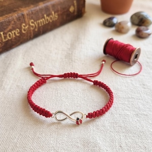 Puede incluir: Una pulsera de cordón rojo con un símbolo de infinito plateado y un pequeño detalle de mariquita roja y negra. La pulsera cuenta con un cierre de nudo corredizo ajustable y está fabricada con hilo rojo trenzado y cuentas plateadas.