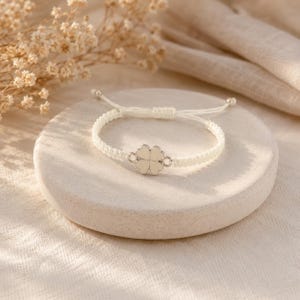 May include: A white macrame bracelet with a four-leaf clover charm. The clover is silver-toned with a white enamel fill. The bracelet is adjustable with sliding knots and small silver beads.