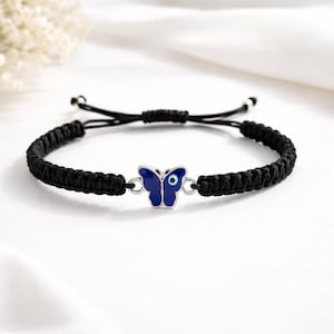 May include: Black braided bracelet featuring a silver butterfly charm with a blue enamel design and an evil eye detail. The bracelet is adjustable with a sliding knot closure. A delicate piece of jewelry.