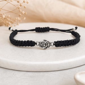 May include: Black macramé bracelet with a silver-coloured rose charm. The adjustable bracelet has a woven design and silver-coloured beads at the ends. A delicate piece of jewellery.