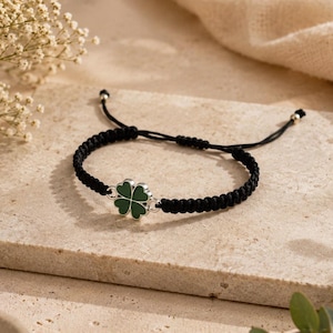 Könnte beinhalten: Ein schwarzes geflochtenes Armband mit einem vierblättrigen Kleeblatt-Anhänger. Das Kleeblatt ist grün mit einem silberfarbenen Rand. Das Armband liegt auf einer strukturierten, hellen Oberfläche mit floralen Akzenten.