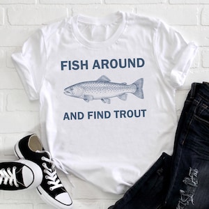 May include: White t-shirt featuring the navy blue text "FISH AROUND AND FIND TROUT" and a trout illustration. The image includes black and white trainers and dark blue jeans.