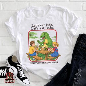 May include: White t-shirt with a cartoon illustration of a green dinosaur serving food to two children. The text reads "Let's eat kids. Let's eat, kids." and "PUNCTUATION SAVES LIVES". The design is set within a red border.