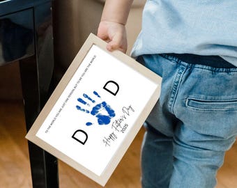 May include: A framed print with a blue handprint and the word "DAD" in black. The text "Happy Father's Day 2025" is below the handprint. The frame is light brown, and the print includes the text "TO THE WORLD YOU ARE JUST ONE PERSON, BUT TO ME YOU ARE THE WORLD."