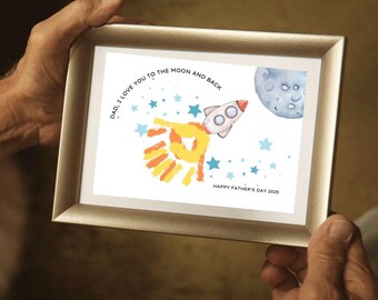 May include: A framed print with a gold-colored frame. The artwork features a rocket ship, a moon, and stars, with the text "DAD, I LOVE YOU TO THE MOON AND BACK" and "HAPPY FATHER'S DAY 2025". The rocket has orange and yellow flames.