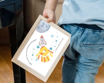 May include: A framed artwork featuring a rocket ship design with a handprint flame, a moon, and stars. The text reads "DAD, I LOVE YOU TO THE MOON AND BACK" and "HAPPY FATHER'S DAY 2023". The frame is light brown.