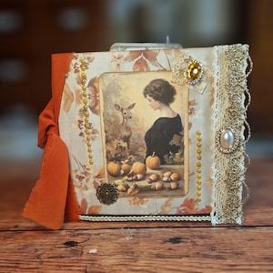 Könnte beinhalten: Ein handgefertigtes, dekoratives Fotoalbum mit Herbstthema. Der Einband zeigt eine Vintage-Illustration einer Frau und eines Hirsches, umgeben von Kürbissen und Pilzen. Verziert mit Spitze, Perlen und einem orangefarbenen Band.