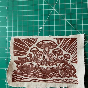 May include: A brown and beige fabric patch featuring a linocut print of mushrooms. The design includes various mushroom shapes, with radiating lines suggesting sunlight. The patch has a raw edge.