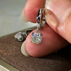 May include: A pair of silver hoop earrings with a dangling clear round gemstone. The earrings are held in a hand.