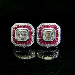 May include: A pair of square-shaped stud earrings. Each earring features a central, square-cut clear stone surrounded by a row of small, red stones. The outer edge is lined with smaller, clear stones. The earrings are set against a black background.