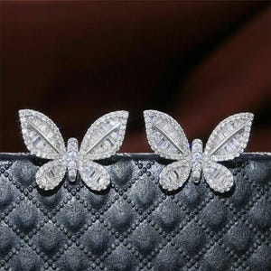 May include: A pair of silver-toned butterfly earrings. Each earring features a butterfly design with encrusted clear crystals. The earrings are set against a black, quilted background.