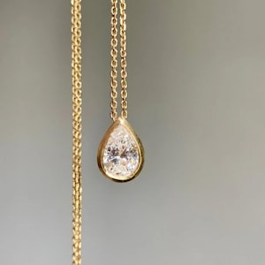 May include: A gold necklace with a pear-shaped pendant. The pendant features a large, clear gemstone set in a gold bezel. The chain is a delicate gold link design, and the background is a solid gray.
