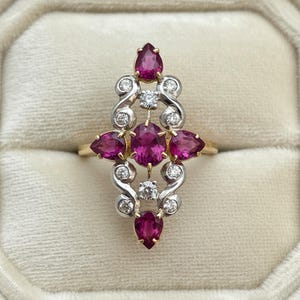 May include: A vintage-style ring featuring a cluster of vibrant, oval and pear-shaped ruby gemstones. The rubies are set in a gold band with silver accents and small, sparkling diamonds. The ring is displayed on a soft, cream-colored surface.