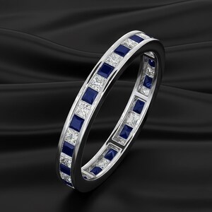 May include: A silver ring with alternating square-cut blue sapphires and clear gemstones. The ring is set against a black background, highlighting the sparkle of the gemstones. The ring is a full eternity band.