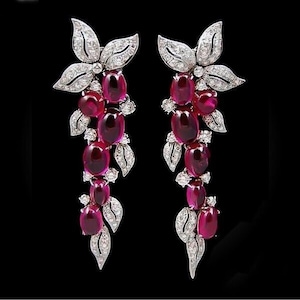 May include: Dangling earrings featuring a floral design with ruby-colored gemstones and small, clear accent stones. The earrings have a silver-colored metal setting and a black background.
