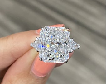 Puede incluir: Un anillo de compromiso de diamante grande de forma cuadrada con tres garras a cada lado. La piedra central es un diamante de corte radiante, y las piedras laterales son triangulares.