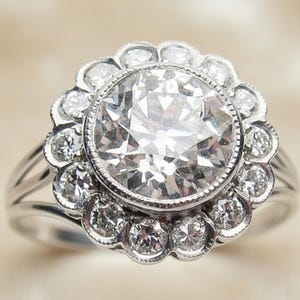 May include: A silver ring featuring a large, round, clear diamond center stone surrounded by smaller diamonds in a floral halo design. The ring has a split shank band.