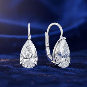 May include: A pair of elegant diamond drop earrings. Each earring features a pear-shaped, clear gemstone set in a silver metal frame. The earrings are displayed against a dark blue, textured background.
