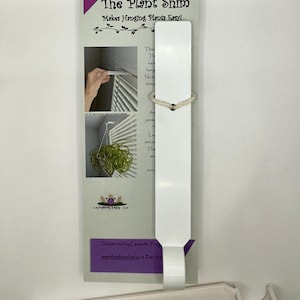 May include: A white plastic plant hanging tool with the text "The Plant Shim" and "Makes Hanging Plants Easy!" printed on a purple and white card. A small, white, rectangular piece is attached to the card with a beige string. A second white piece is at the bottom.