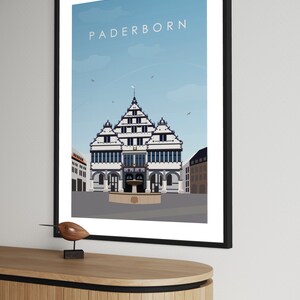 K&ouml;nnte beinhalten: Eine farbenfrohe Illustration des Rathauses in Paderborn, Deutschland. Das Geb&auml;ude ist wei&szlig; mit schwarzen Verzierungen und einem blauen Dach. Der Himmel ist blau mit wei&szlig;en Wolken. Der Text "Paderborn" ist in wei&szlig;en Buchstaben oben im Bild geschrieben.