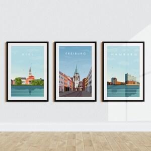 Op de afbeelding: Drie ingelijste stadsgezichten met de namen Kiel, Freiburg en Hamburg gedrukt bovenaan elk frame. De afbeeldingen zijn in een minimalistische stijl met een blauwe lucht en water.