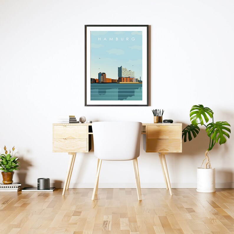 Op de afbeelding: Een ingelijste print van een stadsbeeld met het woord "HAMBURG" bovenaan. Het stadsbeeld is een gestileerde illustratie van gebouwen en water. De print hangt aan een witte muur boven een houten bureau met een witte stoel.