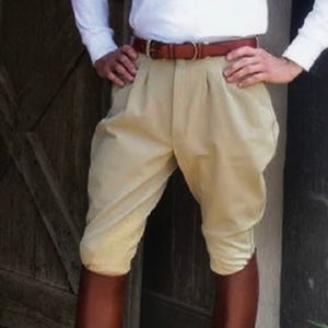 Può includere: Un paio di pantaloni da equitazione beige con una cintura in pelle marrone. I pantaloni hanno un davanti plissettato e sono indossati con stivali da equitazione in pelle marrone.