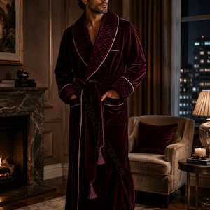 May include: A deep burgundy velvet robe featuring a quilted shawl collar, matching cuffs, and a tie belt with tassel ends. The garment includes thin light-colored piping along the edges and pockets. The text WEDDINGCOLLECTIONHUB is visible.