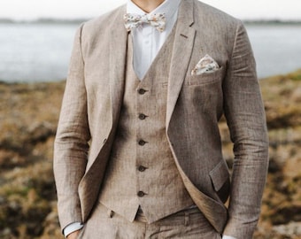 May include: A tan linen suit with a white shirt and a floral bow tie. The suit has a vest and a pocket square.