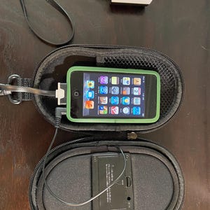 May include: An iPod Touch with a green frame is inside a black zippered case. A black charger cable is connected to the iPod and a white power adapter. A second black case is below the iPod case.