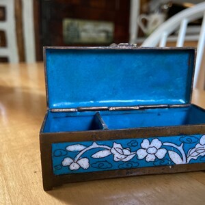 May include: A small, rectangular, brass box with a blue enamel interior and a white floral design on the exterior. The box is open and the lid is propped up.