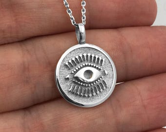 Pode incluir: Um colar com pingente prateado com um berloque circular com um desenho de olho gravado. O olho tem uma íris e cílios detalhados. A corrente do colar também é prateada e tem um delicado padrão de elos. O pingente tem aproximadamente 2,5 cm de diâmetro.