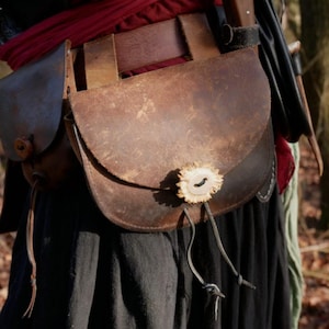 Könnte beinhalten: Eine braune Ledertasche mit abgerundeter Klappe und Geweihverschluss, an einem Gürtel befestigt. Die Tasche hat ein abgenutztes Aussehen, mit einer kleineren Tasche an der Seite. Ein rotes Tuch und schwarze Kleidung sind im Hintergrund zu sehen.