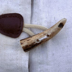 Könnte beinhalten: Nahaufnahme eines Knebelverschlusses. Der Verschluss hat eine braune ovale Lederlasche und einen hellfarbenen Geweihknebel mit zwei Löchern. Der Knebel ist mit einer cremefarbenen Kordel an der Lasche befestigt. Der Verschluss befindet sich auf einem hellfarbenen Stoff.