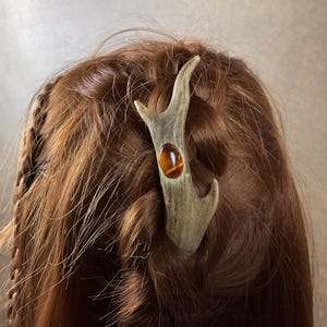 Könnte beinhalten: Ein rustikaler Haarschmuck aus einem Geweihstück, mit einem polierten, ovalen, bernsteinfarbenen Stein. Der Haarschmuck ist in braunem Haar befestigt, mit einem geflochtenen Abschnitt auf der linken Seite. Das Accessoire verleiht einen natürlichen, erdigen Touch.