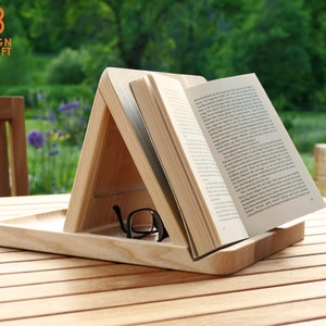 Könnte beinhalten: Ein Holzbuchständer mit einem darauf liegenden Buch. Der Ständer hat eine Ablage darunter, um Gläser oder andere Gegenstände zu halten. Der Ständer steht auf einem Holztisch mit einer Tasse auf dem Tisch.