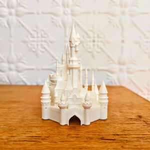 May include: A white miniature castle with multiple turrets, spires, and arched entryways. The model is set on a wooden surface, showcasing intricate architectural details. The castle is a decorative item.