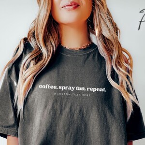 May include: Dark gray t-shirt with the text "coffee. spray tan. repeat." in white. The shirt has short sleeves and a relaxed fit. The text "@CUSTOM TEXT HERE" is below the main text. The model is wearing gray shorts.