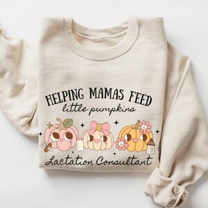 May include: Cream-colored sweatshirt with the text "Helping Mamas Feed Little Pumpkins" above three cartoon pumpkins. The pumpkins are pink, white, and orange, each with unique details. The words "Lactation Consultant" are below the pumpkins.