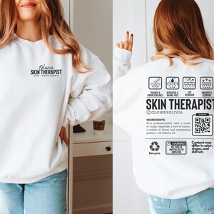 May include: White sweatshirt with the text "Olura SKIN THERAPIST" on the front and a graphic on the back that says "SKIN THERAPIST" with a QR code and the text "GLOWBYOLIVIA".