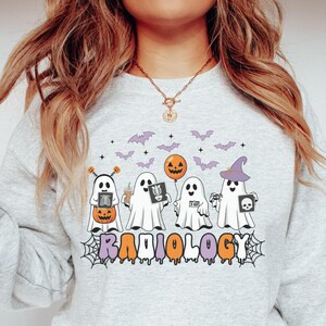 May include: A light gray crewneck sweatshirt featuring a graphic of four ghosts in white sheets. The ghosts hold items including a pumpkin, an orange balloon, and X-ray images. Above them are purple bats and stars. The word RADIOLOGY is printed below.